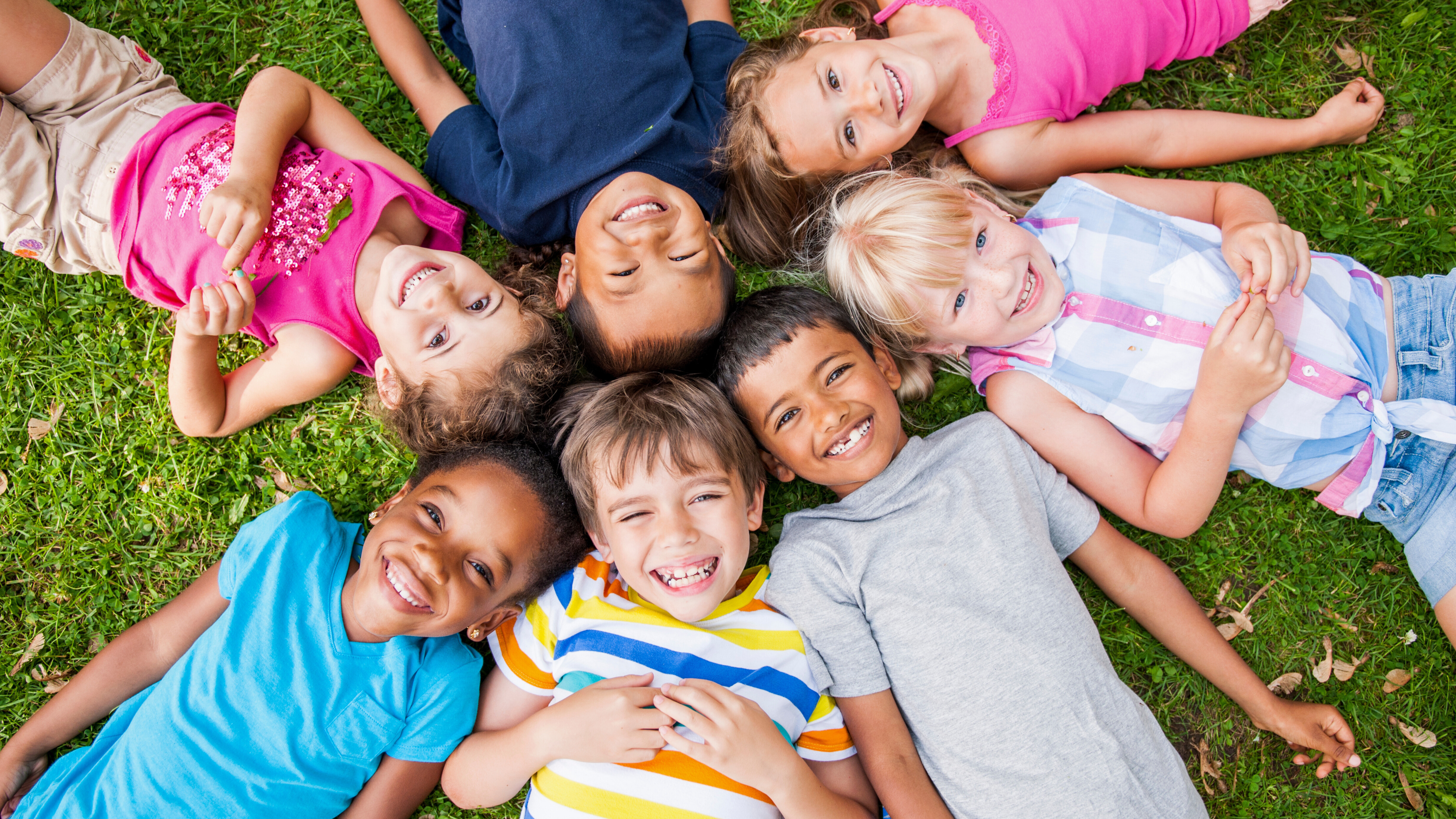 Diverse group of kids laying on the grass in a circle with all the kids smiling. 