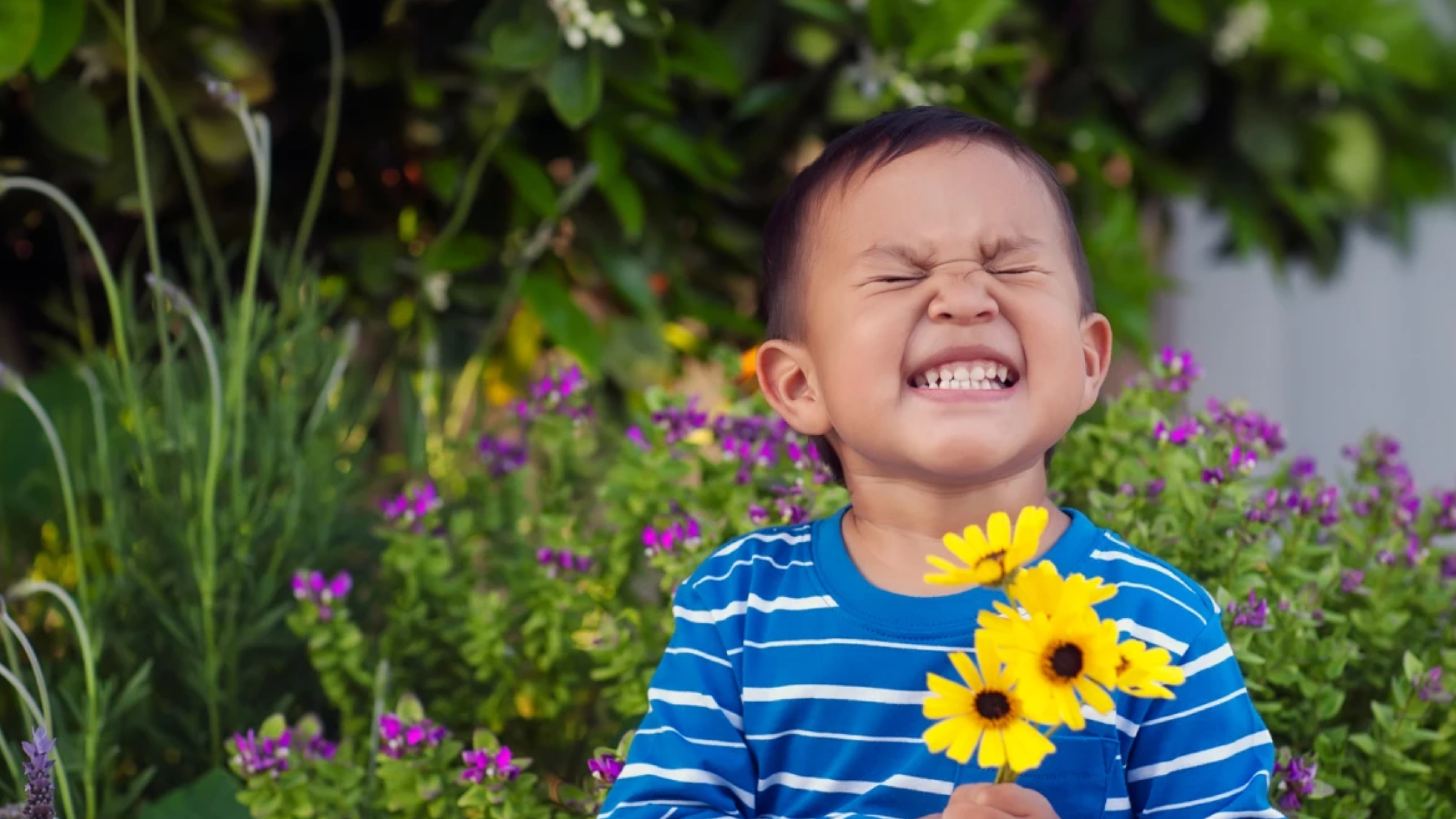 A little boy standing in a garden holding yellow spring flowers smiling very hard.